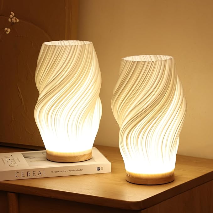 Twee Balaara Serene Wavecrest lampen met golvende patronen, stralen warm wit licht uit op een houten tafel met een boek ernaast, perfect voor een kalme en rustgevende sfeer.