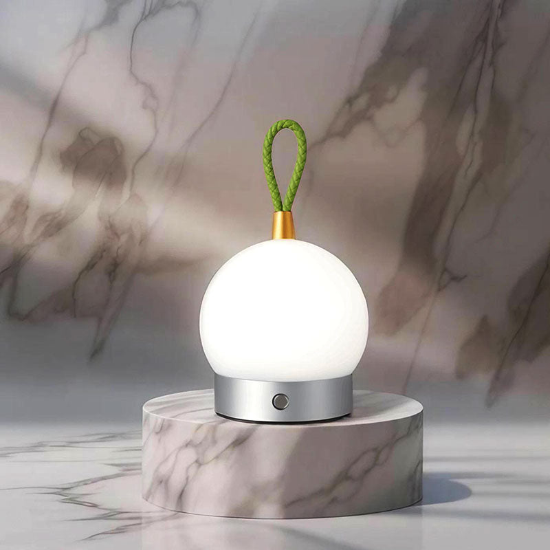 Caleo bedlamp met een modern en minimalistisch ontwerp, bestaande uit een ronde witte bolvormige lamp op een zilverkleurige basis. De lamp heeft een verstelbare groene lus aan de bovenkant voor eenvoudig ophangen of verplaatsen. Geplaatst op een marmeren platform tegen een stijlvolle marmeren achtergrond. Perfect voor een sfeervolle en functionele verlichting in de slaapkamer of woonkamer.