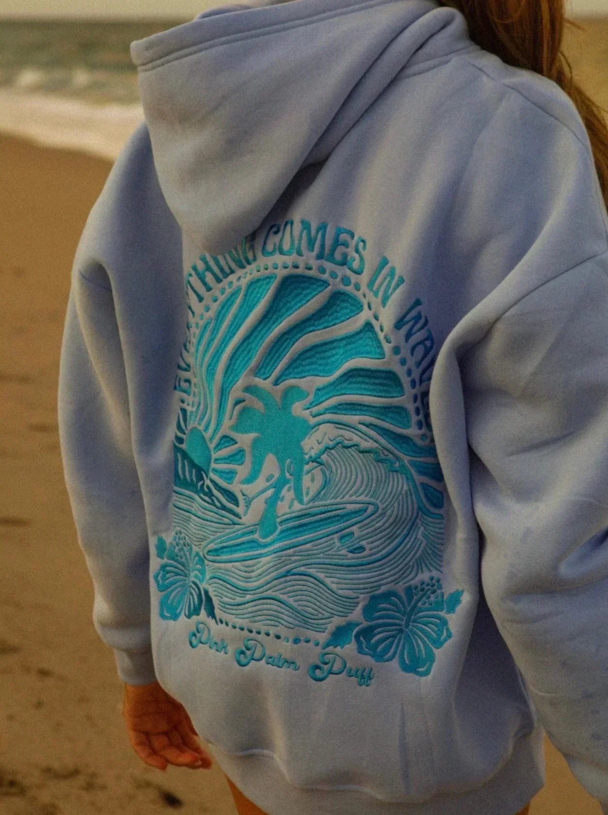 Een vrouw draagt een lichtblauwe hoodie met een opvallende opdruk van een surfsurfer, golven en bloemen op de achterkant. De tekst "EVERYTHING COMES IN WAVES" is zichtbaar, wat de relaxte, surf geïnspireerde stijl benadrukt. De hoodie heeft een comfortabele pasvorm en is perfect voor een casual look aan het strand.