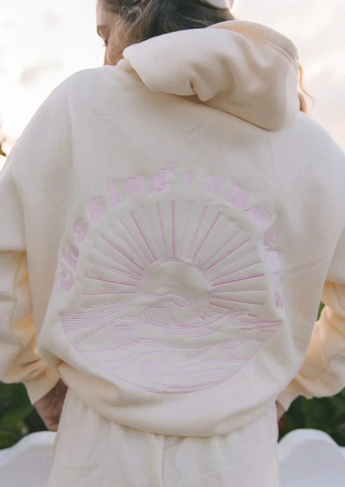 Een vrouw draagt een lichte crème hoodie met de opdruk "chasing sunsets" en een zon met golven op de achterkant. Het zachte ontwerp in pastelkleuren straalt een zomerse sfeer uit, perfect voor ontspannen dagen aan het strand. De hoodie heeft een comfortabele pasvorm, ideaal voor casual looks.