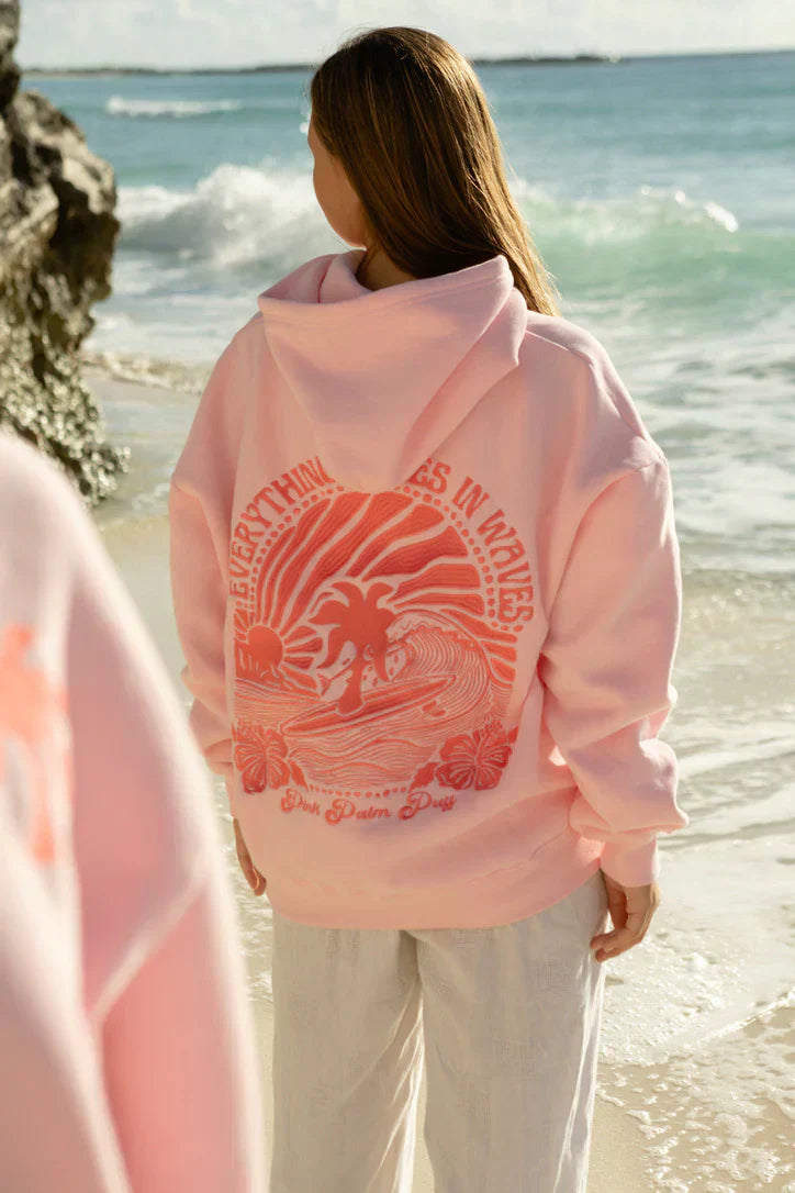 Een vrouw draagt een lichtroze hoodie met een gedurfde, kleurrijke opdruk van een surfer en palmboom op de achterkant, terwijl ze ontspannen langs het strand wandelt. De hoodie heeft een oversized pasvorm en is comfortabel, perfect voor een casual stijl. De tekst op de hoodie luidt